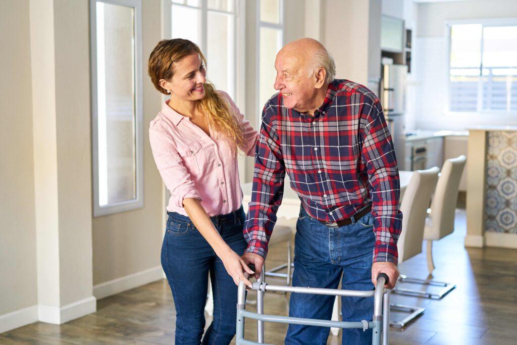 Eine Betreuungsassistentin in einem rosa Hemd hilft einem älteren Mann mit Gehwagen in einem hellen, modernen Wohnraum. Beide lächeln einander an.