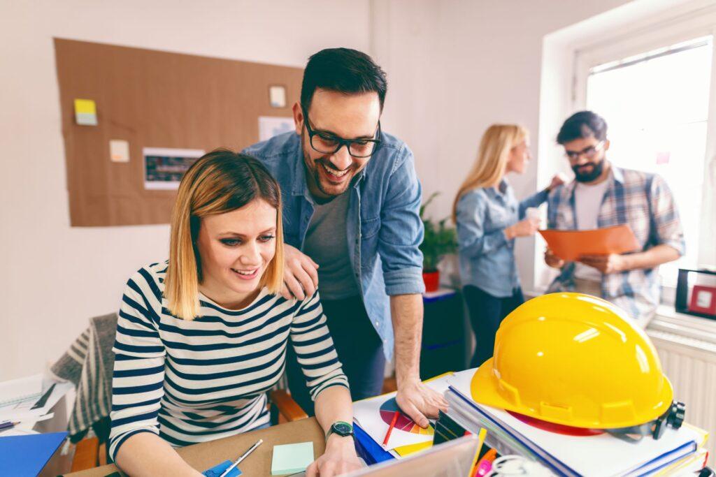 Zwei junge lächelnde Architekten arbeiten an einem großen Projekt. Frau sitzt und verwendet Laptop, während Mann mit Brille dahinter steht und schaut, was sie tut. Start up Business-Konzept.