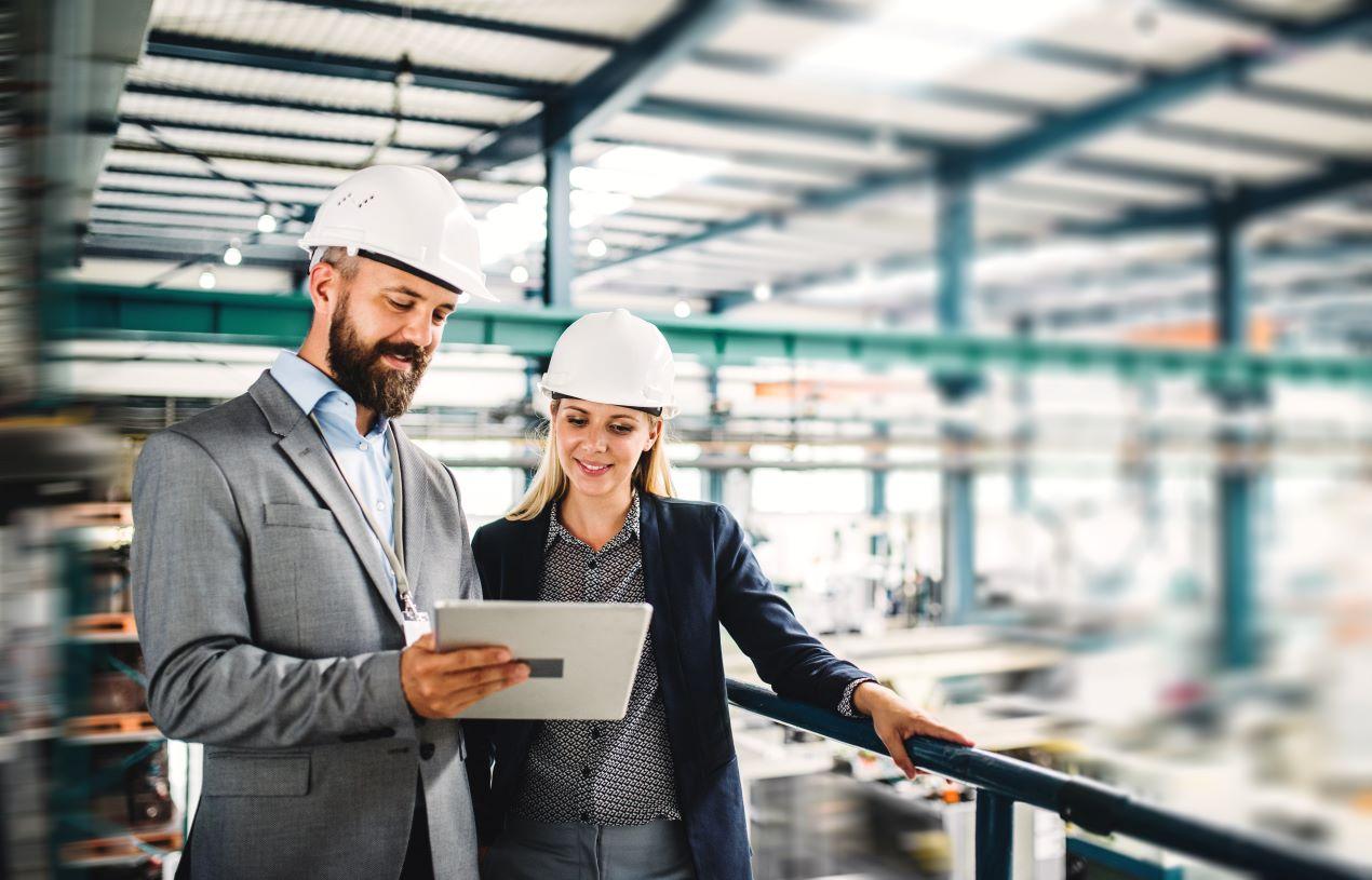 Zwei Ingenieure mit Helmen analysieren Bodenmarkierungskonzepte in einer modernen Industriehalle zur Verbesserung der Sicherheitsstandards.